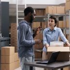 Warehouse team discussing about distribution logistics while putting client goods in carton box after wrapping order in bubble wrap. Industrial employee working with merchandise in storehouse