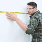 Male carpenter measuging white wall with measure tape at home