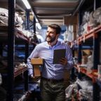 Smiling bearded businessman walking trough storage of shipping firm, holding boxes under armpit and using tablet to check on goods.