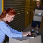 Pretty lady is using a laptop in a warehouse, next to a gray-haired man with boxes