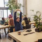 Businessman is stretching his arms above his head while sitting at his desk in a modern coworking space, surrounded by colleagues working on their laptops