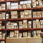 African American warehouse worker checking inventory next to large racks of cardboard boxes holding product in a distribution warehouse.