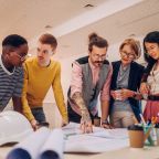 Portrait of a diverse team of architects using blueprints in the office. Focused multiracial engineers cooperating to create new building project. Focus on a tattooed man boss holding a meeting.
